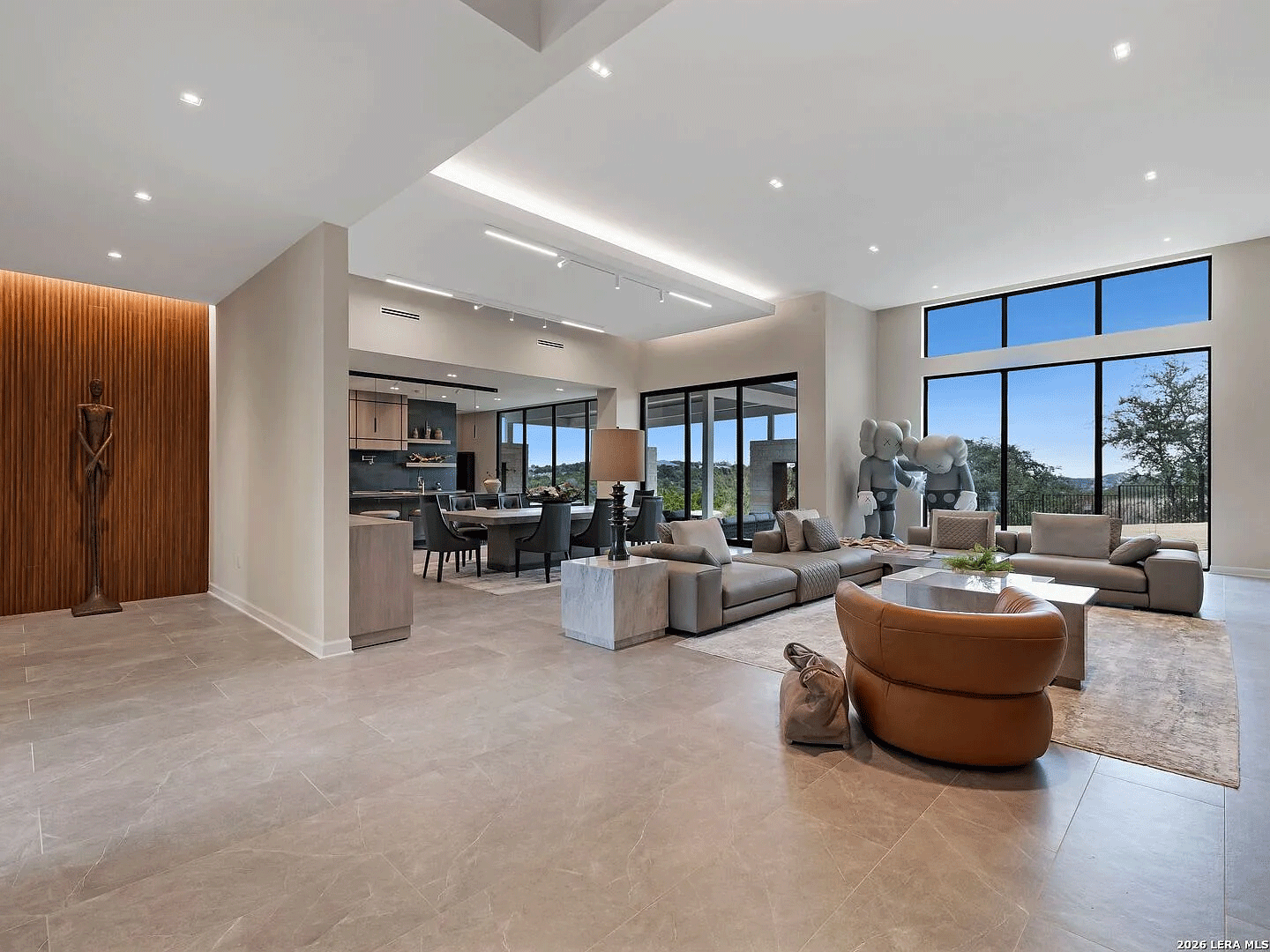 Luxury Interior Design in Texas by Khamila Design House featuring a furnished open-concept living room in a modern Texas Hill Country home. Custom sectional sofa, sculptural lounge chair, stone flooring, warm wood accent wall, and integrated architectural lighting with expansive windows overlooking scenic views in San Antonio luxury home.
